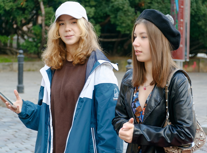 13 - Two young friends standing outdoors in an urban area, with the person on the left wearing a blue-and-white jacket, and the person on the right in a black beret and leather jacket